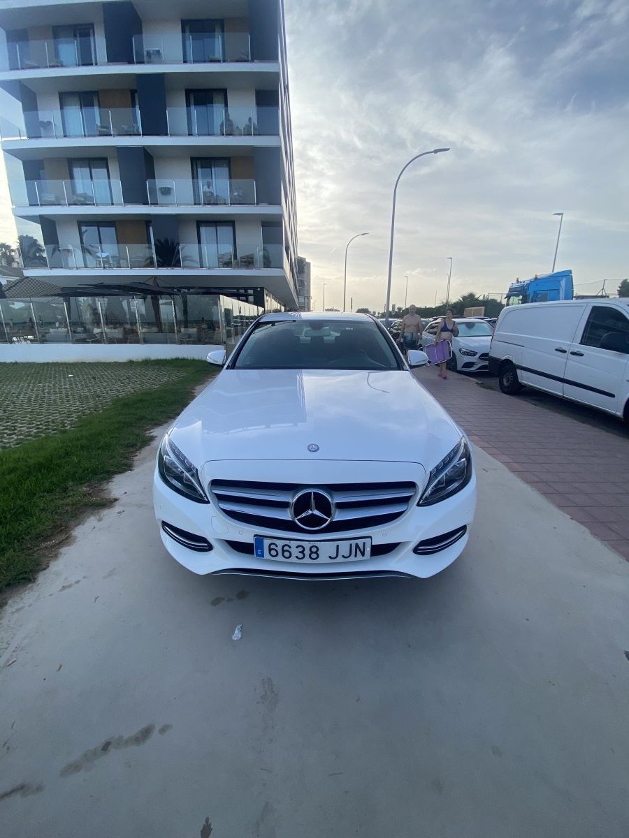 Un sedán Mercedes blanco estacionado en una acera junto a un edificio de apartamentos al anochecer.