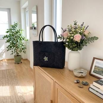 Eine schwarze Tragetasche auf einem Holzschrank in einem hellen Flur, umgeben von Blumen und einem gerahmten Foto.