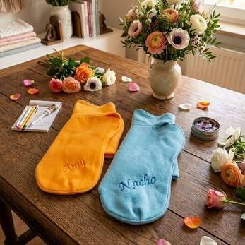 Zwei personalisierte Hundepullover, orange und blau, auf einem Holztisch mit Blumen und Bastelmaterialien
