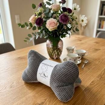 Ein graues, gestricktes, hundeförmiges Kissen liegt auf einem Holztisch, im Hintergrund steht eine Blumenvase.