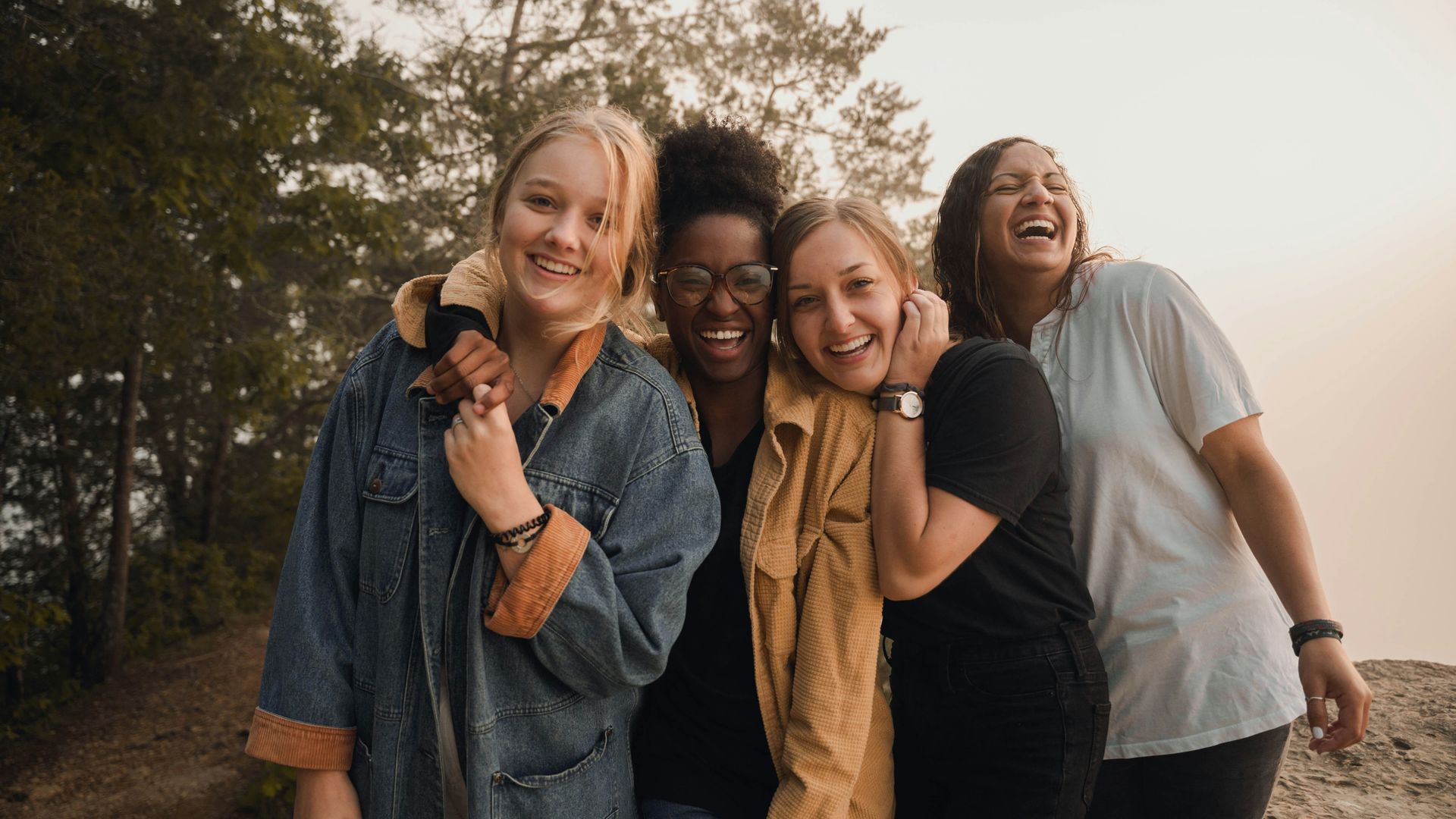 Cuatro mujeres diversas riendo y abrazándose al aire libre cerca de los árboles con la cálida luz del sol.