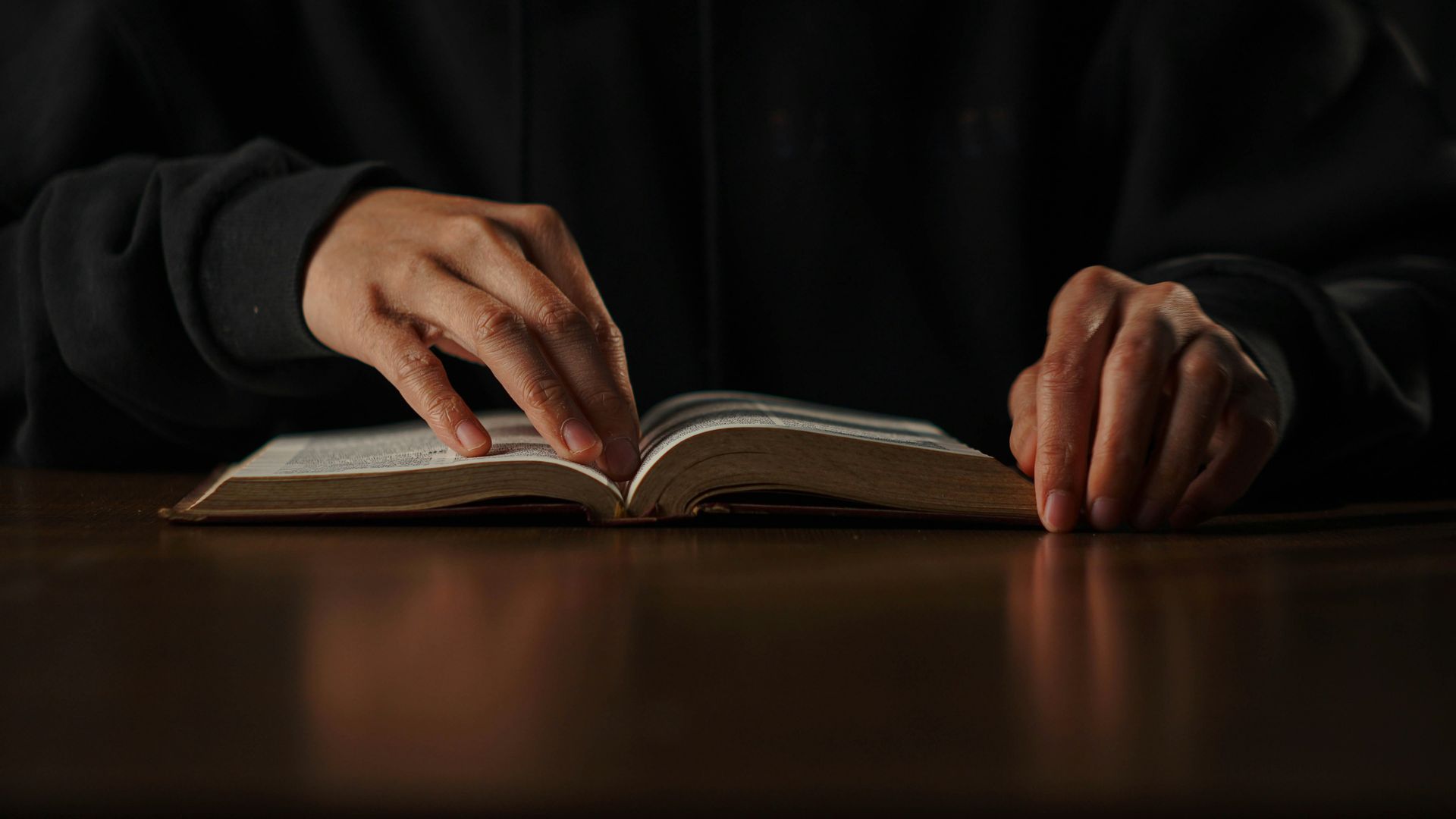 Una persona con sudadera negra lee un libro abierto sobre una mesa de madera oscura. Sus manos pasan las páginas.