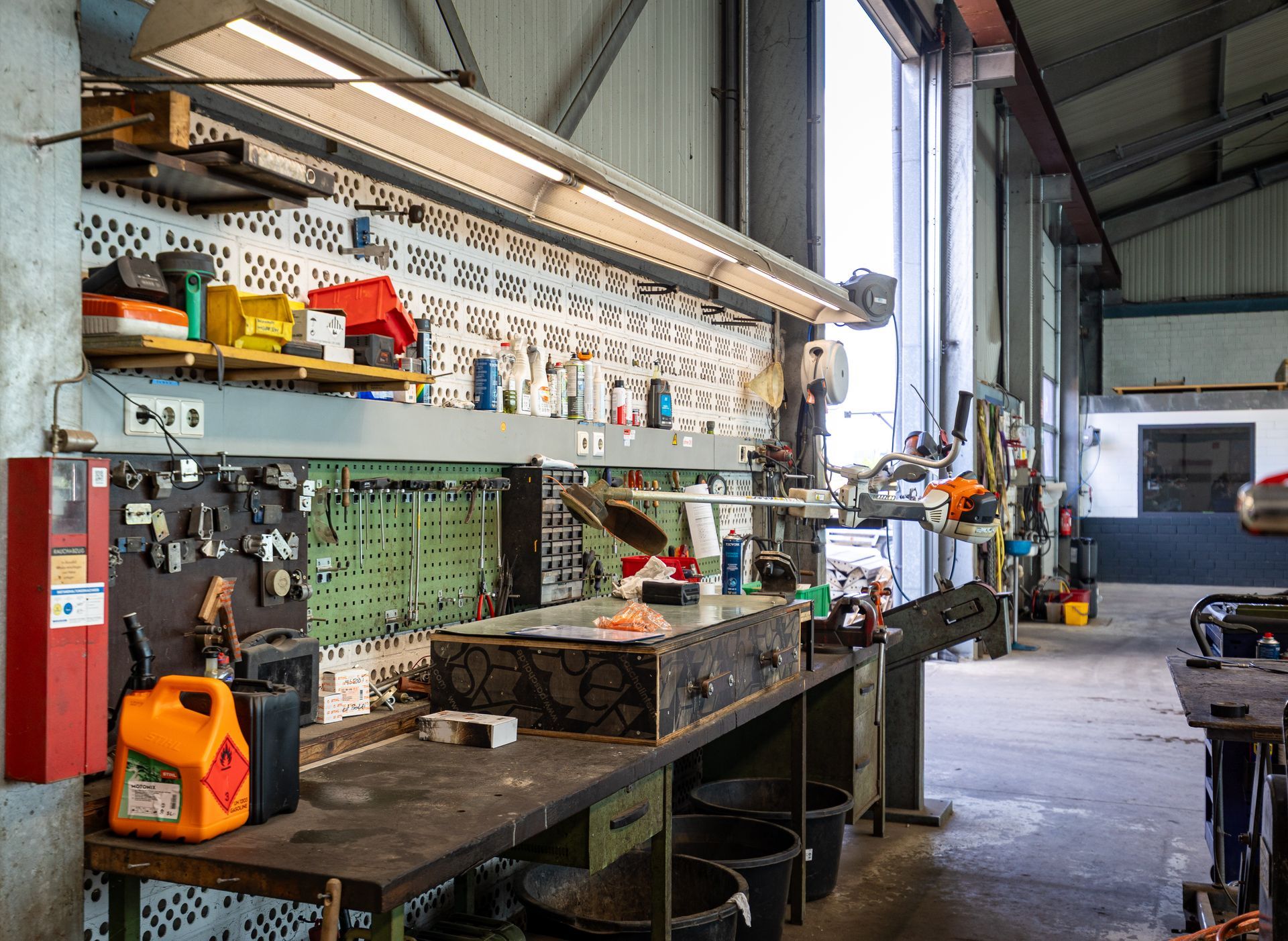 Werkstattinterieur mit Werkbank, Werkzeugen, Regalen und leuchtend orangefarbenen Geräten in der Nähe einer großen, offenen Tür
