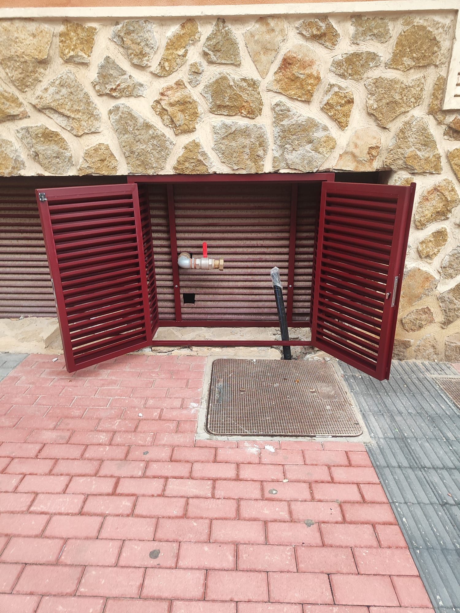 Una puerta roja está abierta en una acera de ladrillo frente a un muro de piedra.