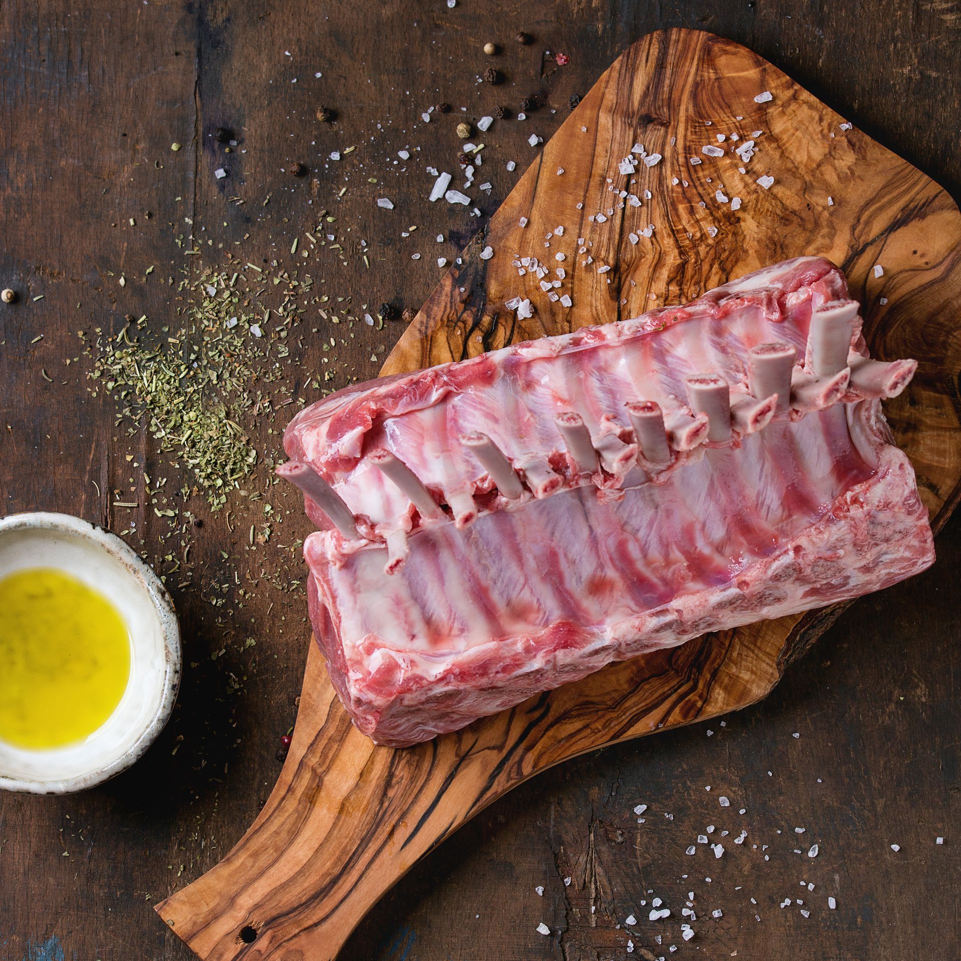 Carré de côtes crues sur une planche à découper en bois avec du gros sel et un bol d'huile sur une table sombre