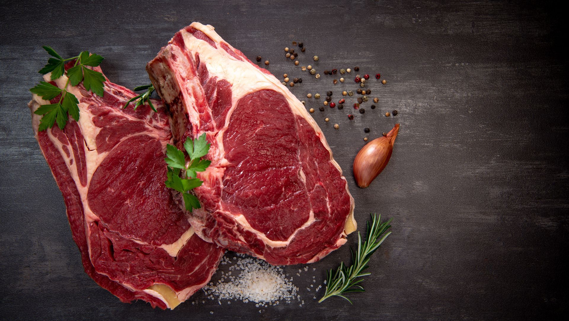 Deux steaks de bœuf crus avec des herbes, des grains de poivre et de l'ail sur une surface d'ardoise sombre