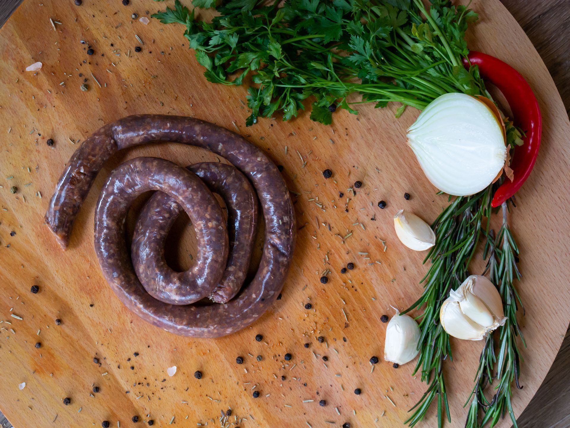 Saucisse crue enroulée sur une planche en bois avec des herbes, de l'ail, de l'oignon, du piment et des grains de poivre.