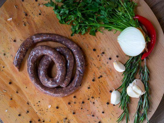 Saucisse crue enroulée sur une planche en bois avec des herbes, de l'ail, de l'oignon, du piment et des grains de poivre.