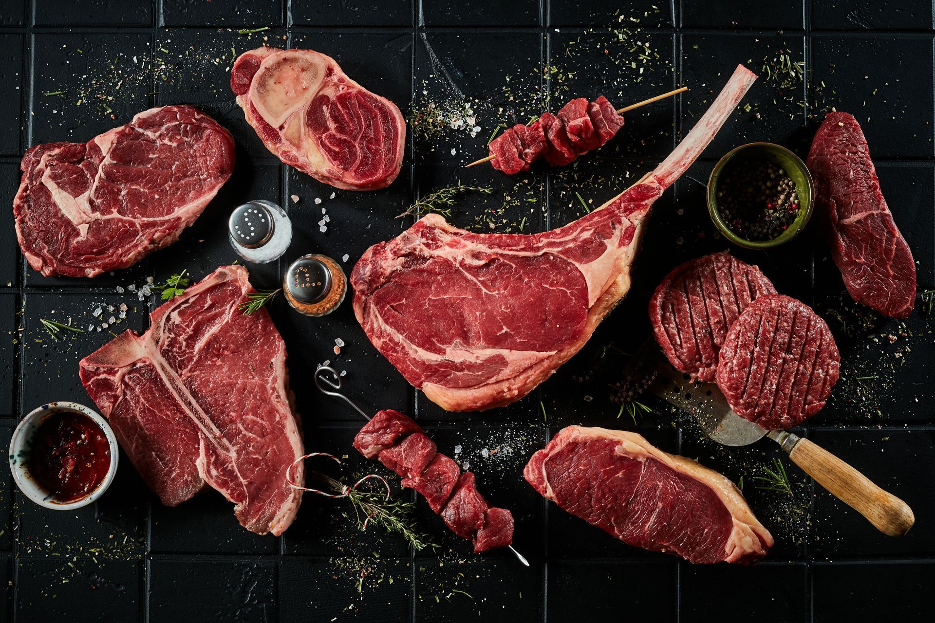 Assortiment de steaks et de morceaux de bœuf crus disposés sur une ardoise sombre avec des herbes et des épices.