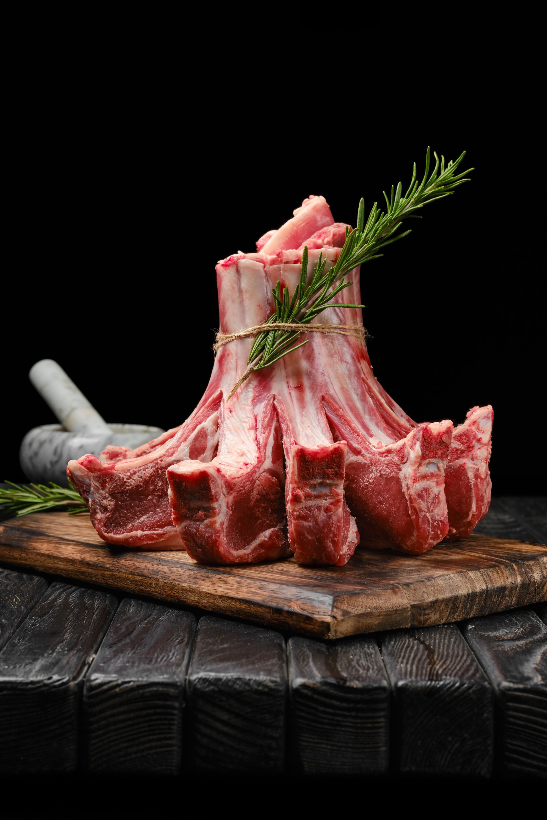 Carré d'agneau cru sur une planche en bois, garni de romarin et de poivre sur fond sombre