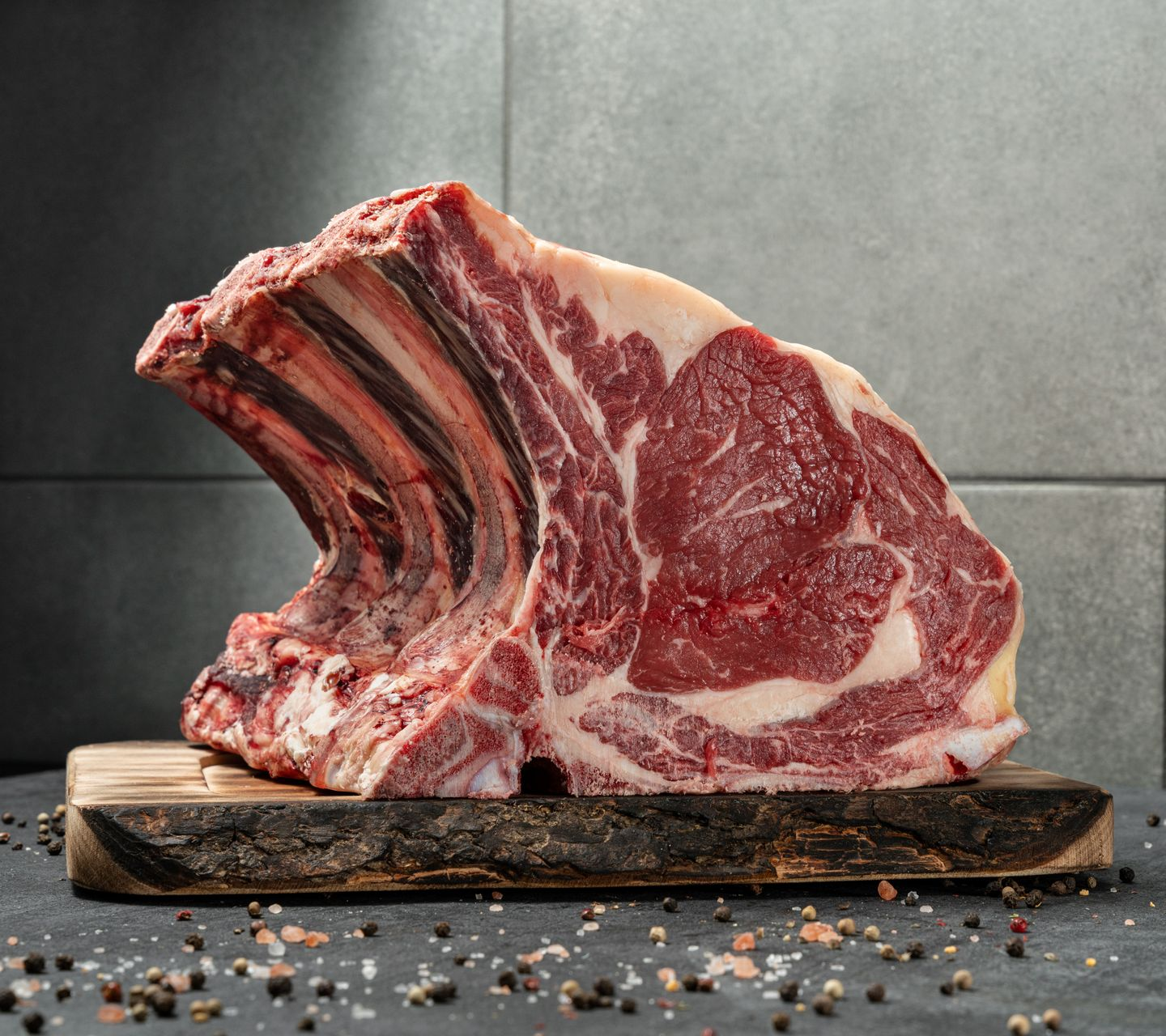 Steak tomahawk cru sur une planche en bois, avec os et gras persillé visibles, sur une surface en pierre sombre.
