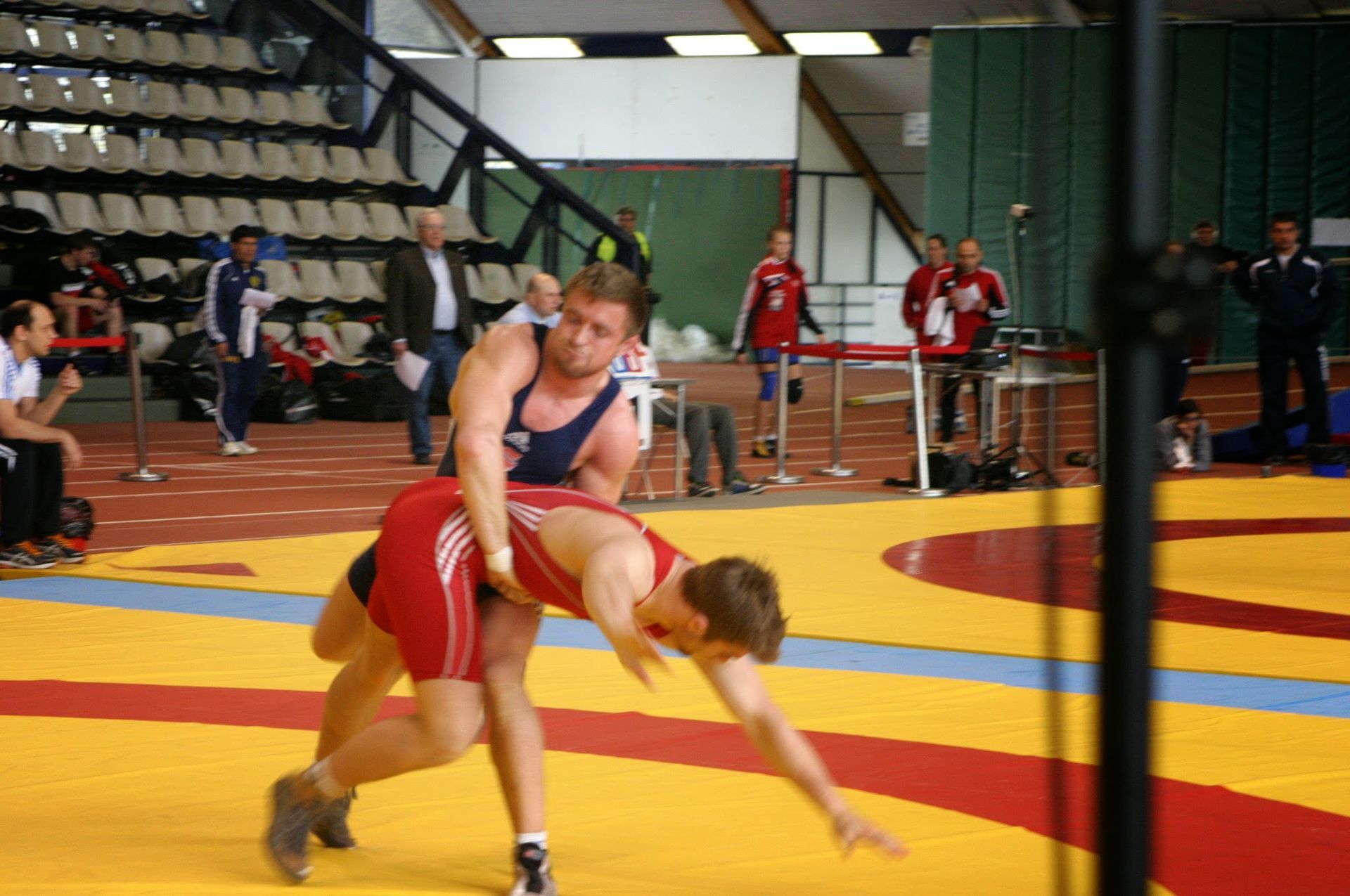 Kaksi painijaa painii keltaisella ja punaisella matolla kilpailullisessa ottelussa sisäurheiluareenalla.