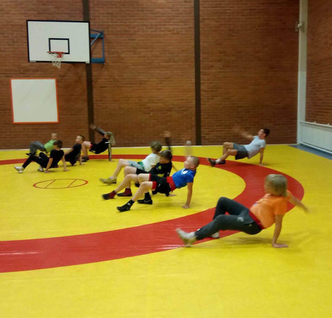 Ryhmä ihmisiä suorittaa rapukävelyjä keltaisella ja punaisella painimatolla tiiliseinäisessä liikuntasalissa.