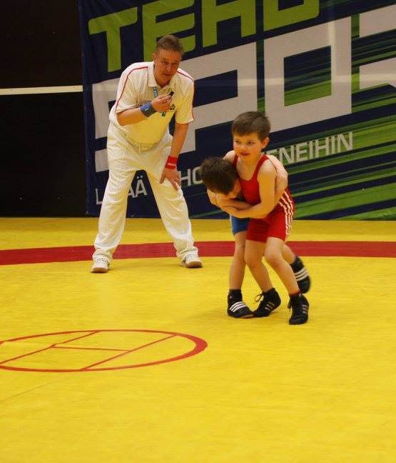 Tuomari katsoo, kun kaksi nuorta painijaa punaisissa ja sinisissä painijapaidoissa ottavat yhteen keltaisella matolla sisäareenalla.