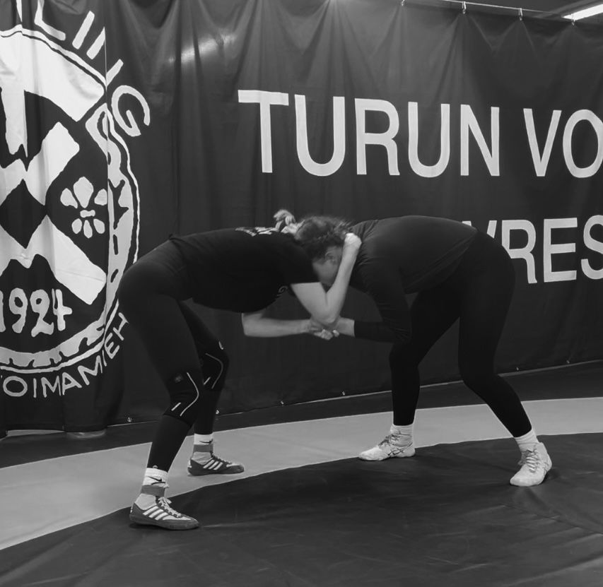 Kaksi urheiluvarusteisiin pukeutunutta ihmistä painii painiasennossa matolla Turun Voimamiehet -banderollin edessä.
