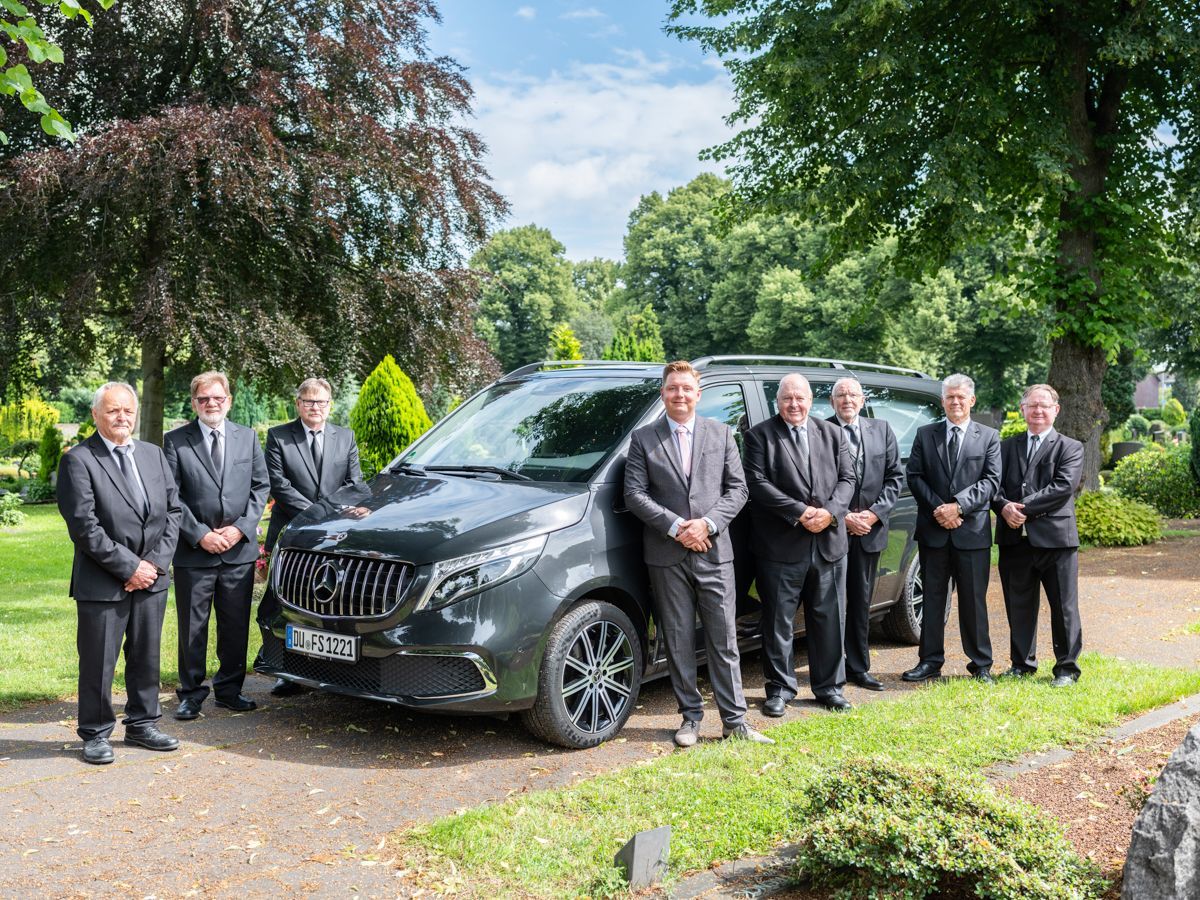 Eine Gruppe von Männern in Anzügen steht vor einem schwarzen Leichenwagen.