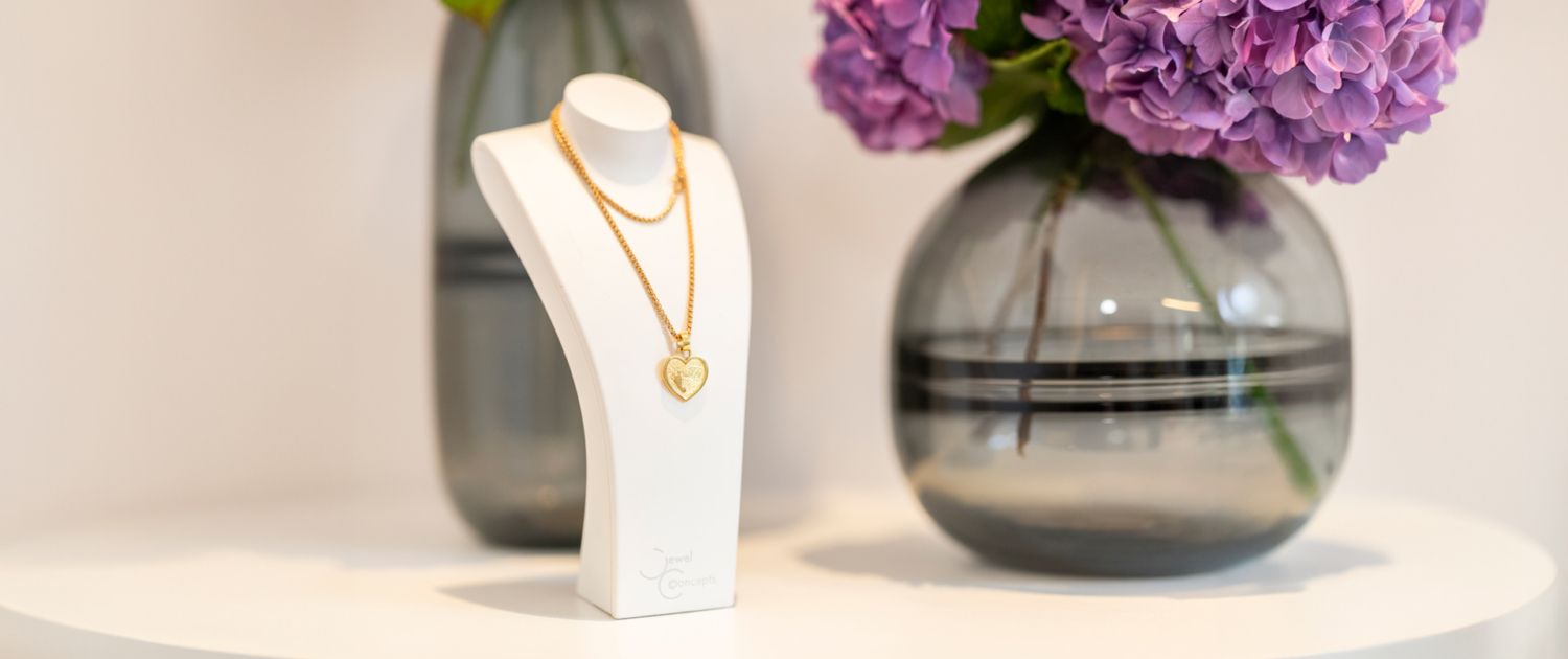 Eine Schaufensterpuppe trägt eine Halskette auf einem Tisch neben einer Vase mit lila Blumen.