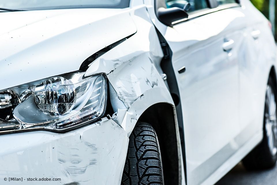 Beschädigtes weißes Auto; Kotflügel und Tür vorne verbeult. Scheinwerfer und Reifen sichtbar.