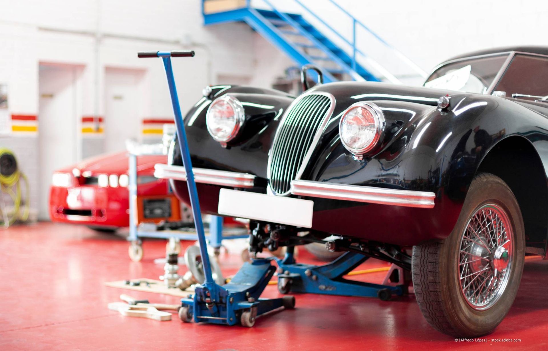 Ein schwarzer Oldtimer steht in einer Garage auf einem Wagenheber, im Hintergrund ist ein rotes Auto zu sehen.