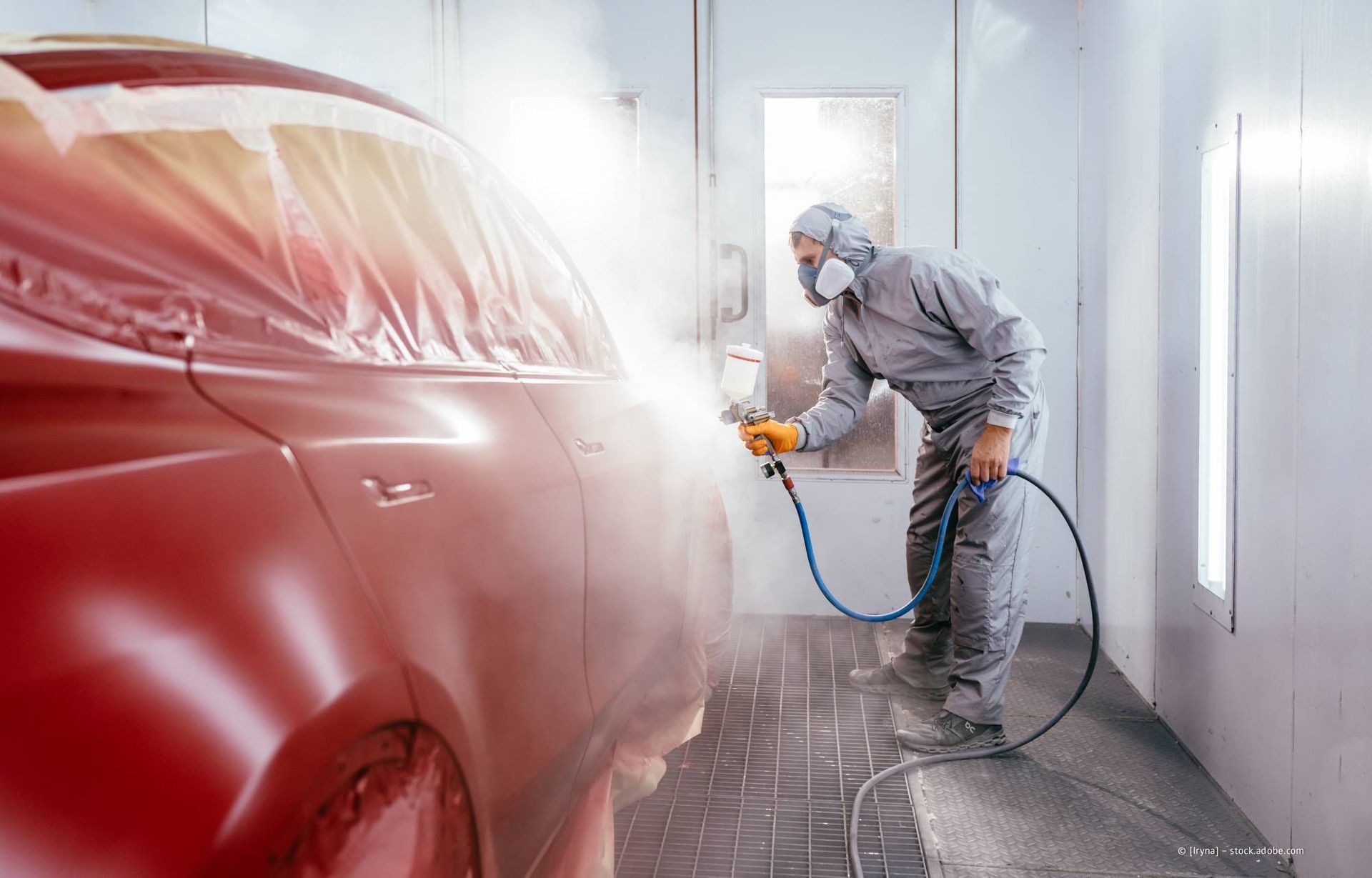 Eine Person in Schutzkleidung besprüht ein Auto in einer Lackierkabine mit roter Farbe.