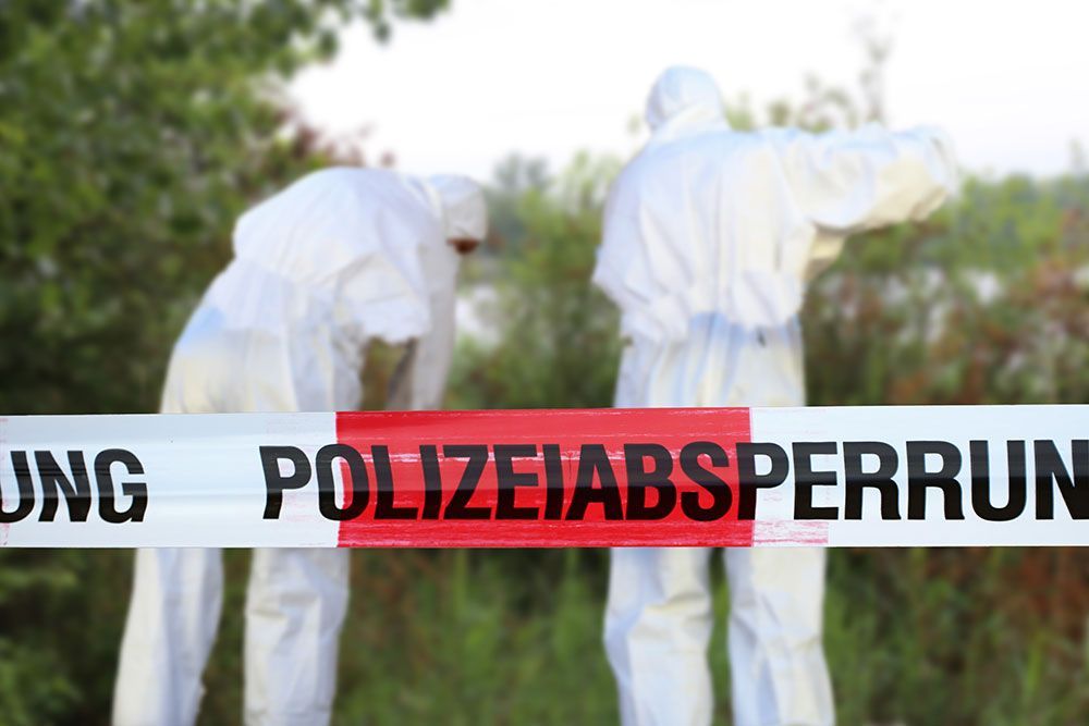 Zwei Forensiker in weißen Schutzanzügen hinter Polizeiabsperrband in einem grasbewachsenen Außenbereich