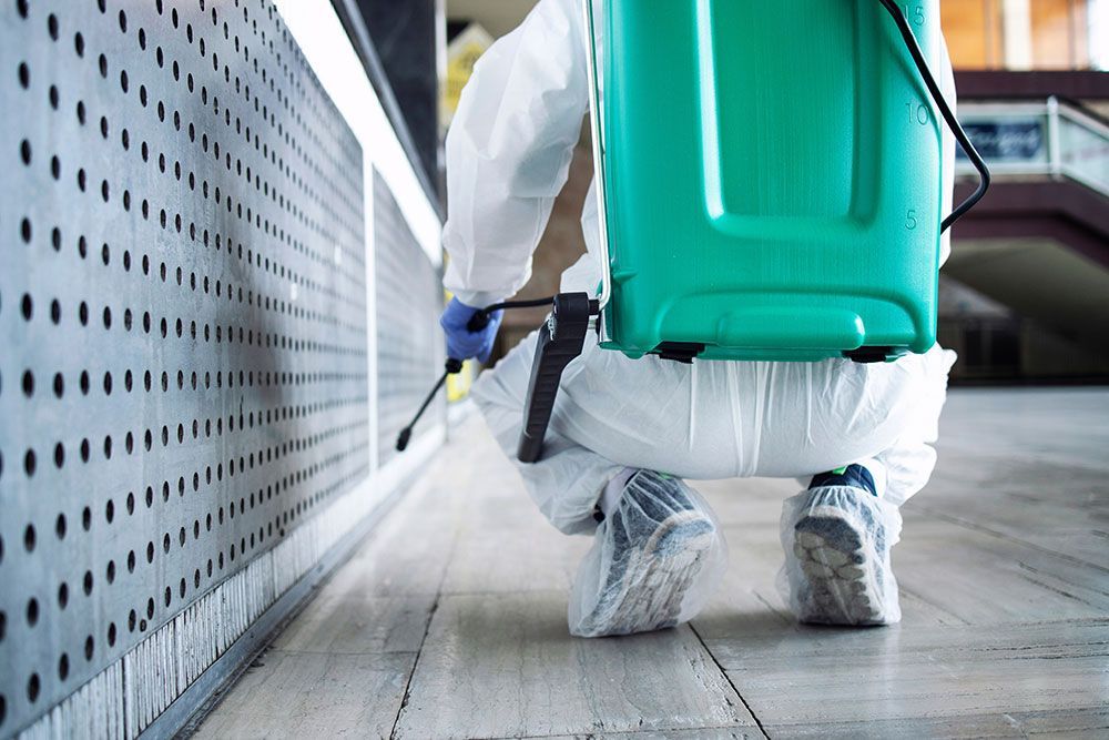 Ein Arbeiter in weißer Schutzkleidung sprüht Desinfektionsmittel neben eine Gebäudewand.