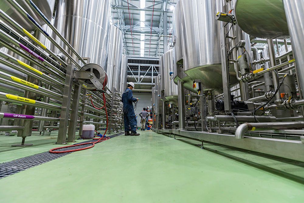 Innenansicht einer Industrieanlage mit großen Edelstahltanks, Rohren und Arbeitern auf einem grünen Bodengang.