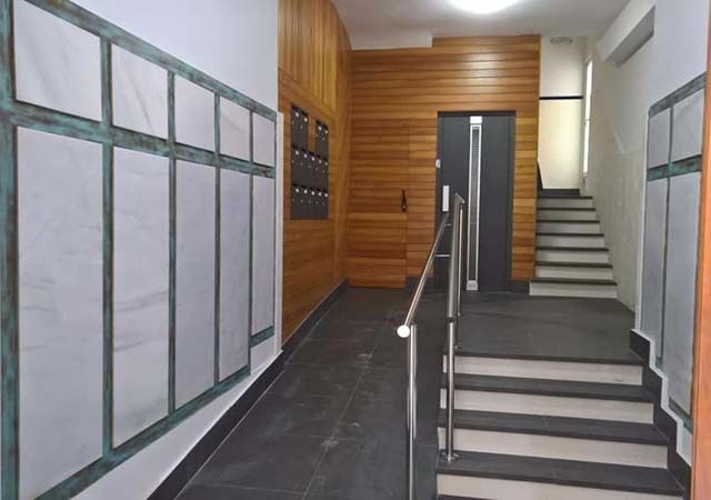 Escalera interior con peldaños grises, pasamanos metálicos y una pared revestida de madera en el descansillo.
