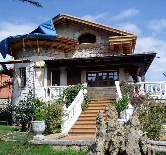 Casa de piedra con barandillas blancas y escaleras de madera, vista desde un jardín bajo un toldo azul.