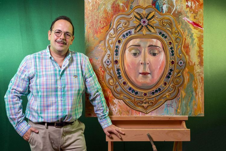 Una persona se encuentra de pie junto a un cuadro que representa un rostro estilizado y ornamentado, enmarcado en un marco enjoyado, sobre un fondo verde.