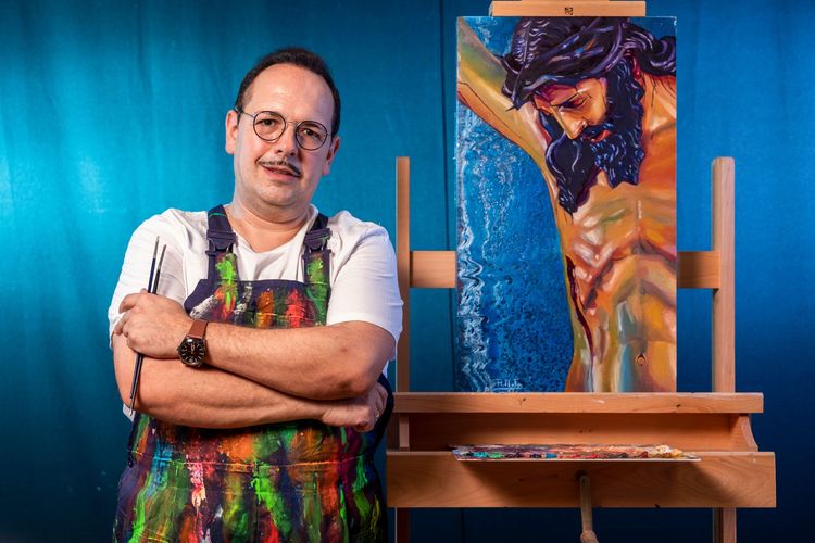 Un artista con gafas y delantal permanece de pie con los brazos cruzados junto a un caballete donde se exhibe un cuadro de una figura crucificada.