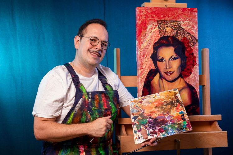 Un artista con un delantal salpicado de pintura sostiene una paleta junto a un retrato pintado sobre un caballete con fondo azul.