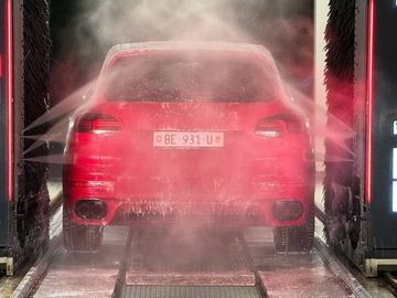 Ein rotes Auto wird in einer automatischen Waschanlage gereinigt, wobei Wasserstrahlen das Fahrzeug von den Seiten besprühen.