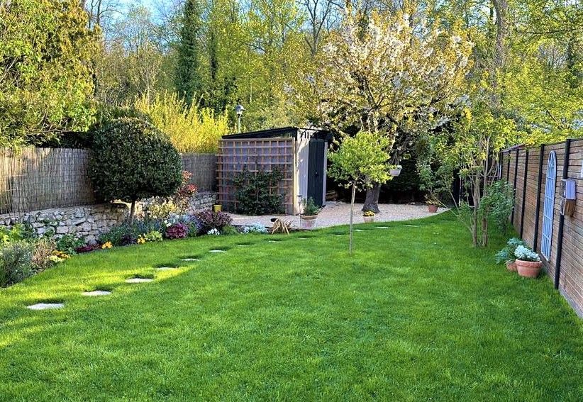Pelouse verte dans un jardin ensoleillé avec allée en pierre, arbres, abri de jardin et clôture