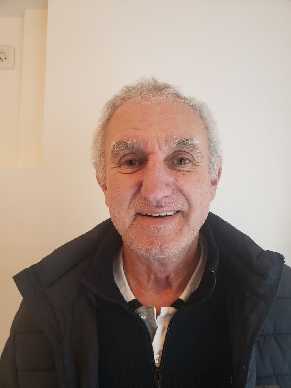 Un homme souriant aux cheveux et à la barbe grisonnants, vêtu d'un costume claire et d'une chemise blanche