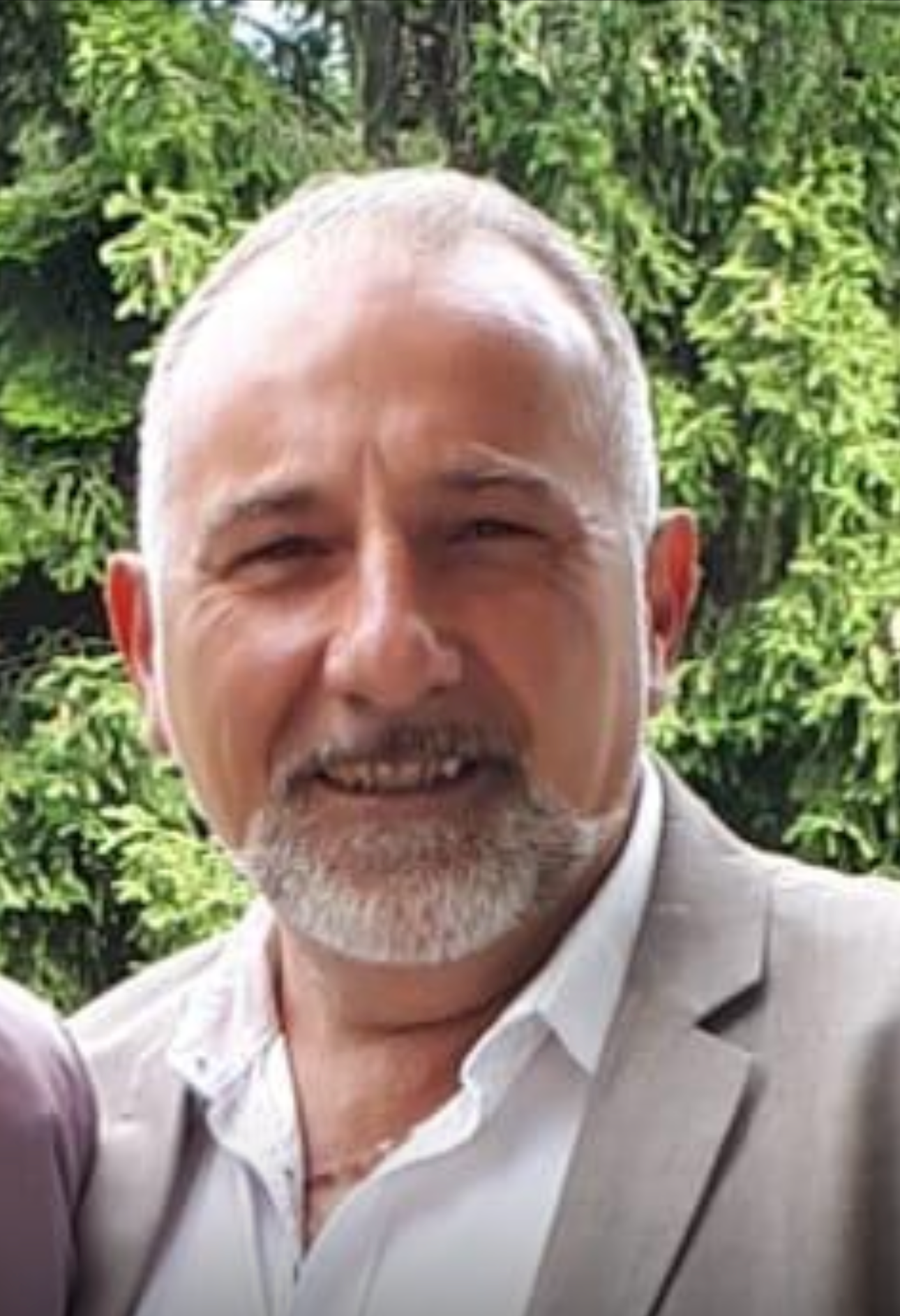 Un homme souriant aux cheveux et à la barbe grisonnants, vêtu d'un costume claire et d'une chemise blanche