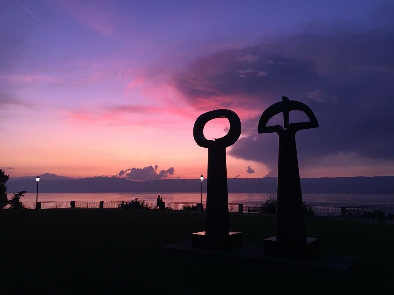 Silhouette de deux sculptures se détachant sur un coucher de soleil aux nuances de rose, de violet et de bleu sur l'eau.
