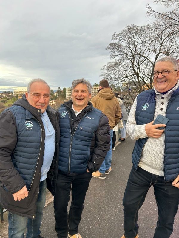 Trois hommes souriants en extérieur, vêtus de gilets et de vestes bleus, vue de la ville en arrière-plan.