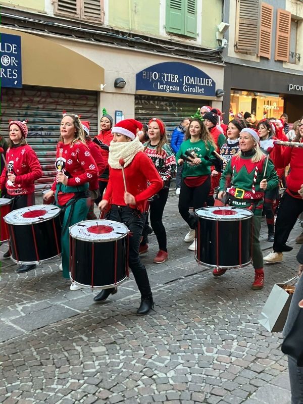 Des personnes en pulls de Noël défilent et jouent du tambour dans une rue.