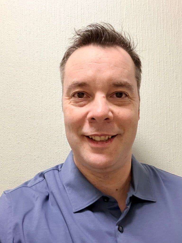 Un homme souriant aux cheveux et à la barbe grisonnants, vêtu d'un costume claire et d'une chemise blanche