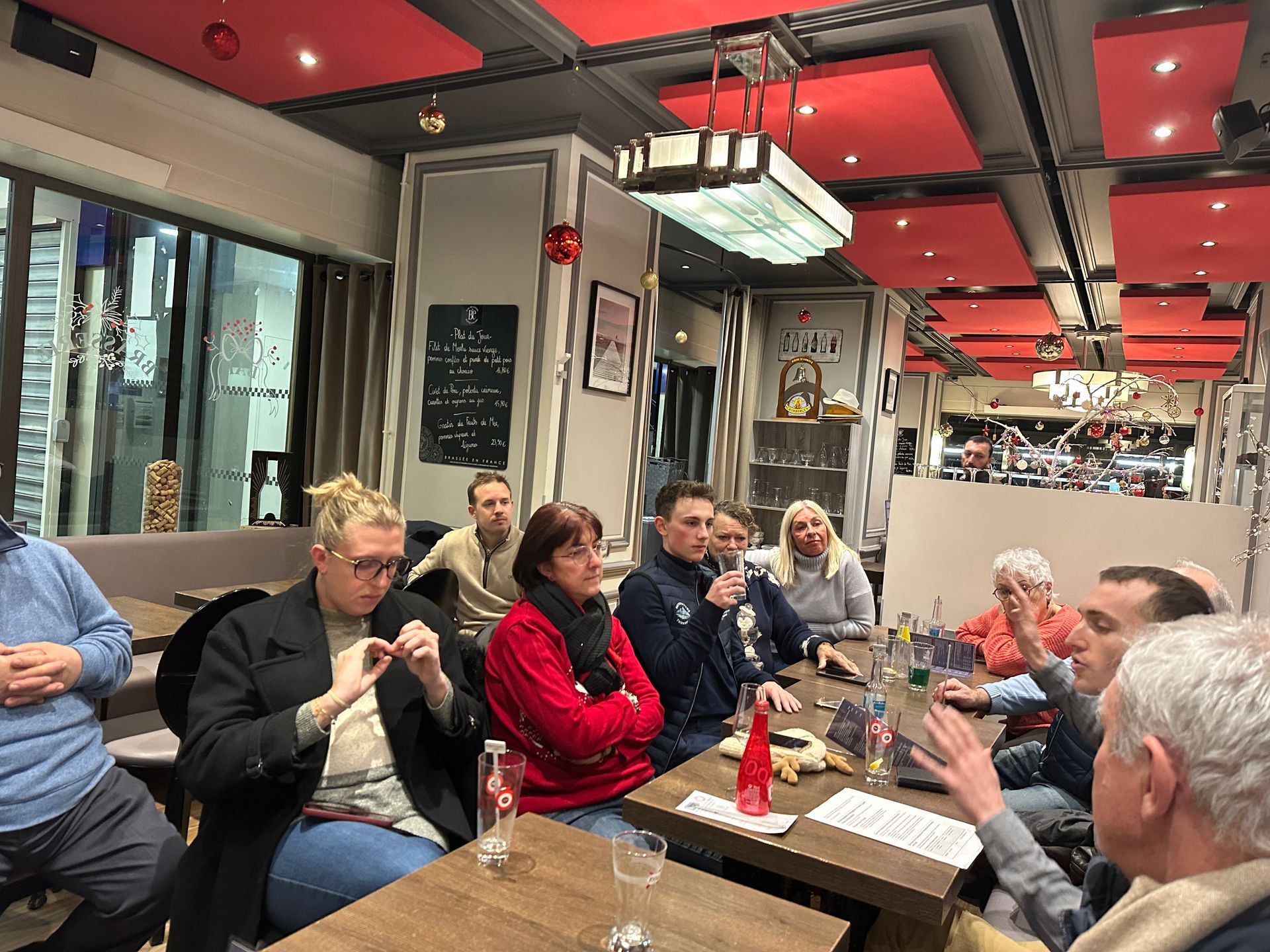 Des personnes sont assises autour d'une table dans un restaurant. Un homme parle. D'autres écoutent tenant un verre.