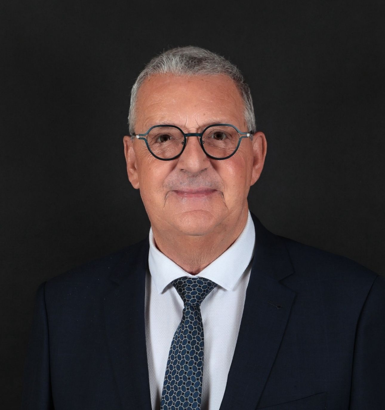 Homme en costume et lunettes, souriant, sur fond sombre.