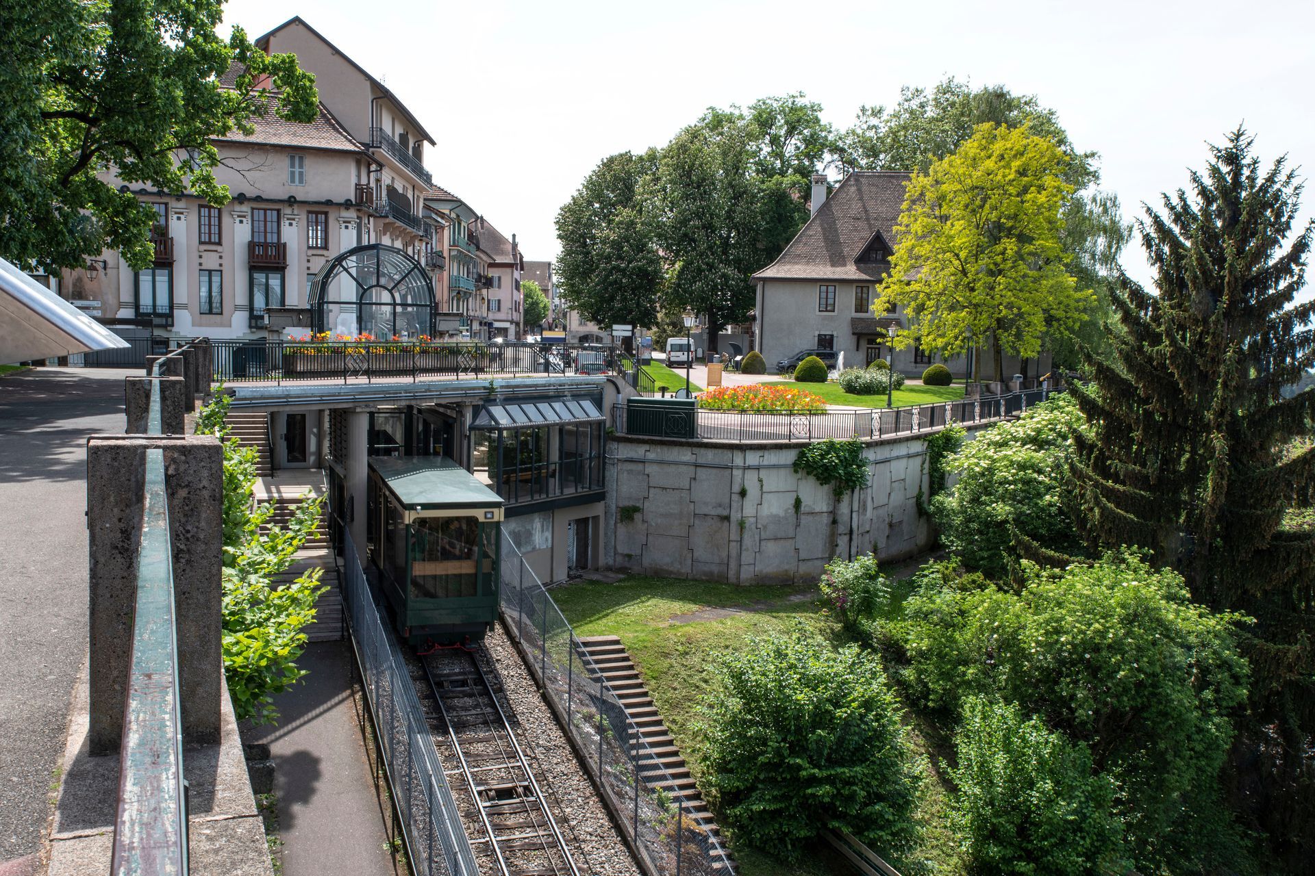 Un funiculaire gravit une voie escarpée sur une place de la ville. Train vert, bâtiments en pierre et végétation luxuriante.