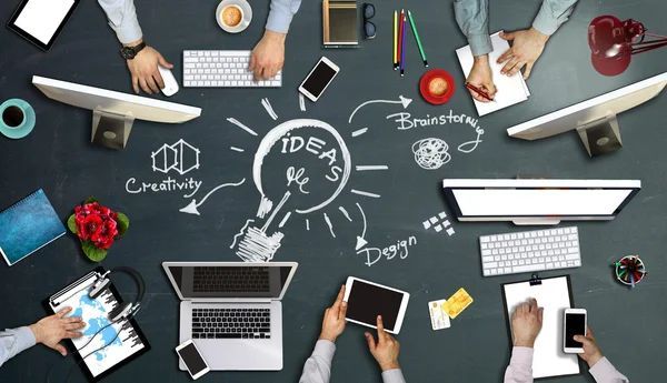 Trabajo en equipo alrededor de un escritorio con computadoras portátiles, tabletas, café y un boceto en una pizarra con la etiqueta