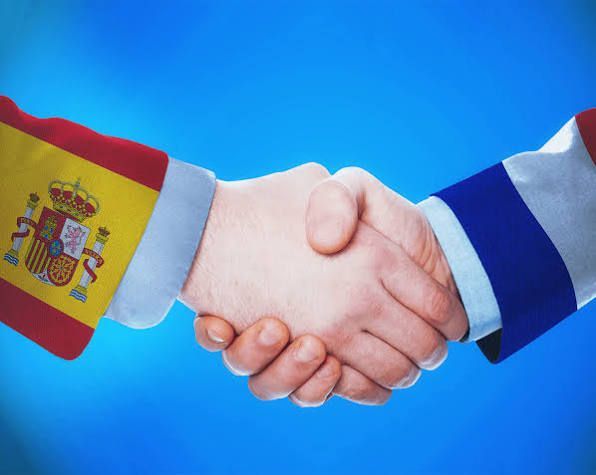 Saludo de manos entre dos personas, una con la manga de la bandera española y la otra con la manga azul y blanca, sobre un fondo azul.