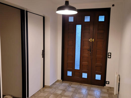 Un hall d'entrée lumineux avec un sol carrelé, une porte blanche sur la gauche et une porte d'entrée en bois foncé.