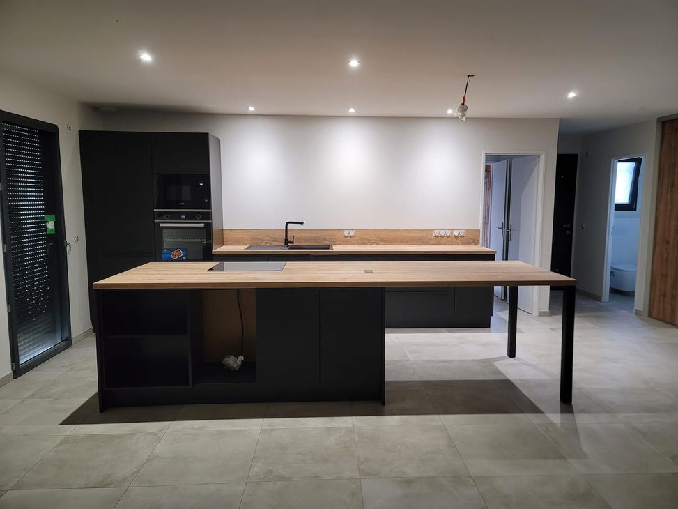 Cuisine moderne avec îlot et placards noirs, plans de travail en bois clair et carrelage neutre dans une pièce spacieuse.