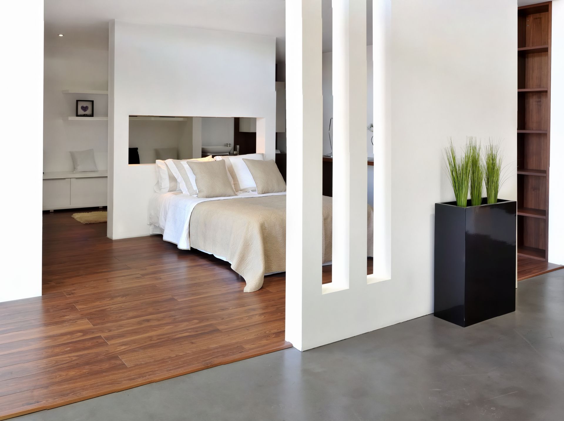 Une chambre moderne avec un lit aux tons neutres, visible à travers une découpe dans une cloison blanche.