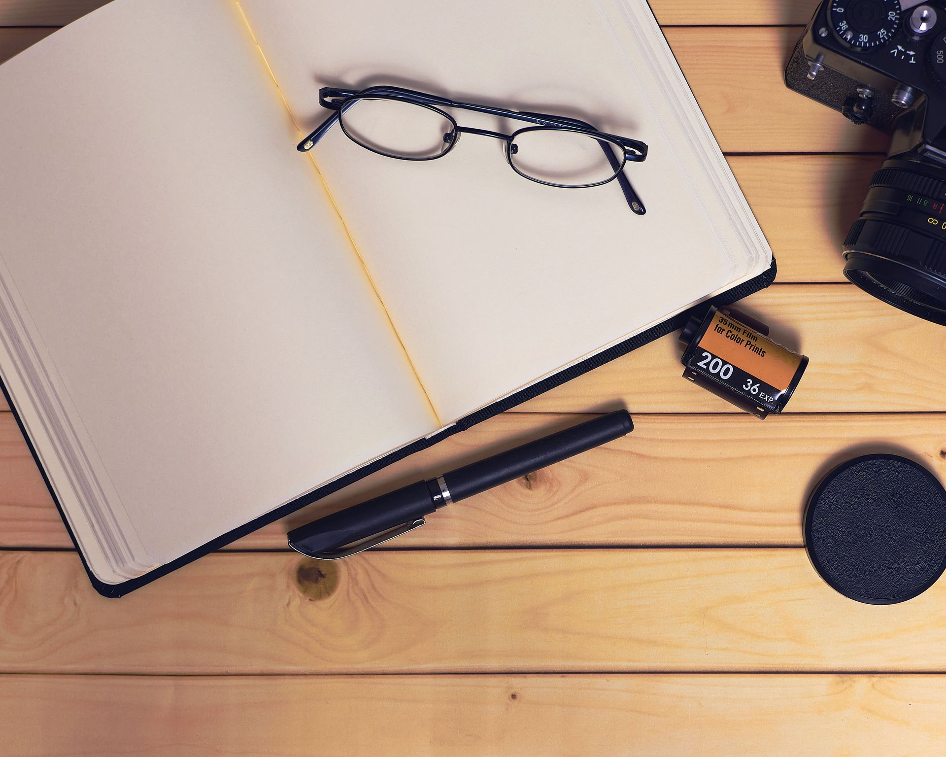 Un cuaderno abierto con gafas, un bolígrafo y una cámara sobre una mesa de madera.