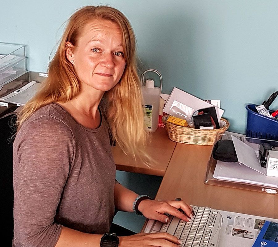 Frau mit langen blonden Haaren tippt an einem Computer an einem Schreibtisch. Blaue Wand im Hintergrund.